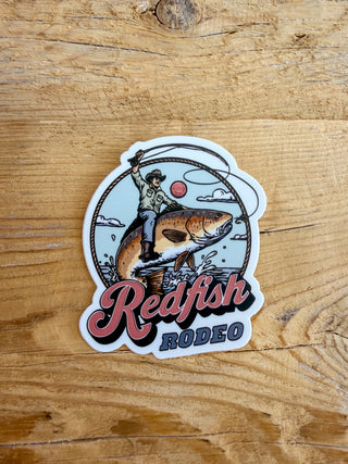 A sticker with a graphic of a cowboy riding a redfish with a fly fishing rod and text that says Redfish Rodeo