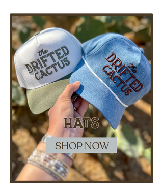 Two hats with 'The Drifted Cactus' branding held by a hand against a natural background.
