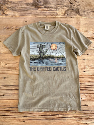 Khaki comfort colors unisex fit tee with a cactus growing on the coastline graphic, sun and sky in the background. 