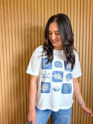 White boxy unisex fit tee with a blue checkered design with seashell, cactus, and sun elements. 