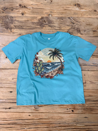 A turquoise blue short-sleeve t-shirt with a circular graphic print of a beach scene featuring a palm tree, cacti, and waves, and fall leaves to make it a coastal fall tee. 