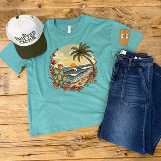 A turquoise blue short-sleeve t-shirt with a circular graphic print of a beach scene featuring a palm tree, cacti, and waves, and fall leaves to make it a coastal fall tee. 
